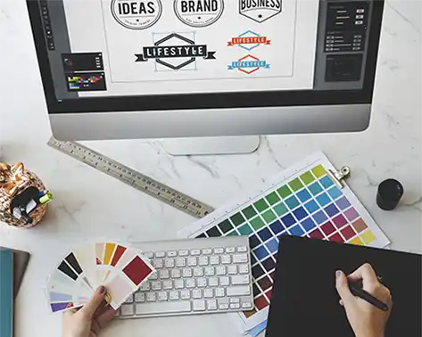 Graphic design workspace with color swatches, keyboard, and computer monitor displaying logo designs.